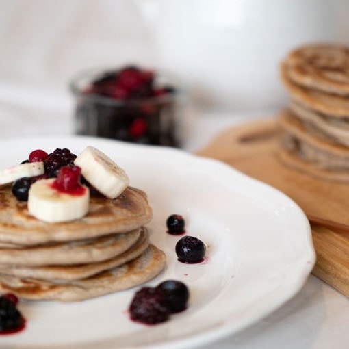 A stack of pancakes topped with banana slices and berries on a white plate, with more berries in a jar and another stack in the background.