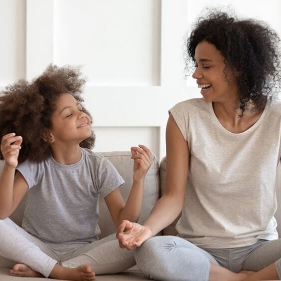 Mother and daughter sitting cross-legged on a couch, smiling at each other, wearing casual gray outfits in a bright room.