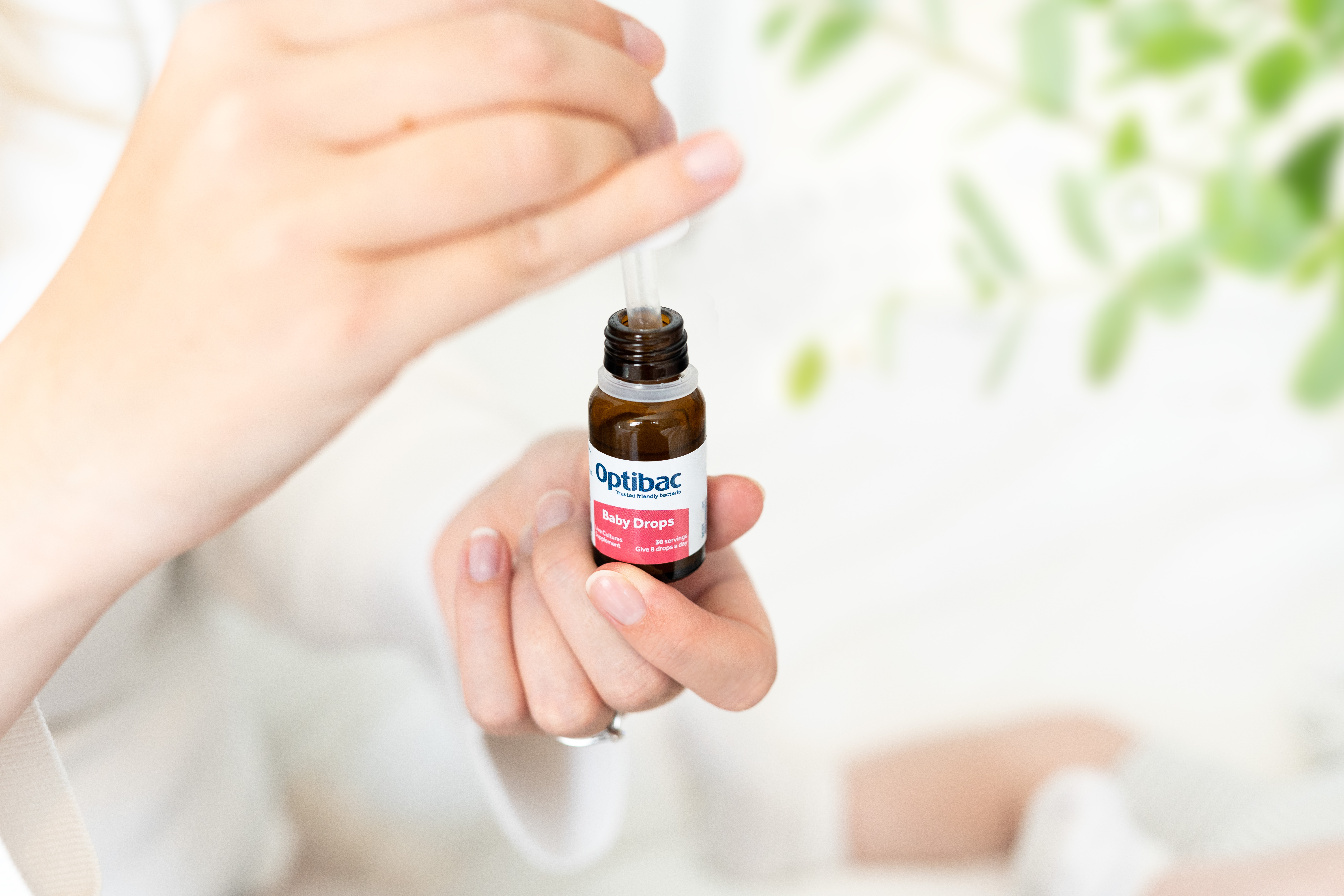 A person holding a small bottle of Optibac Baby Drops, using a dropper, with a blurred leafy background.