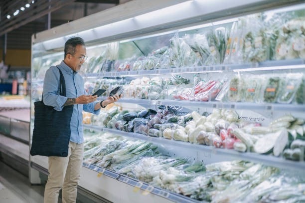 Man shopping in a grocery store, holding a phone and looking at packaged vegetables on a refrigerated shelf.