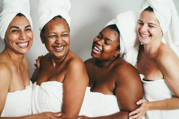 Four women wrapped in towels and with towels on their heads, smiling and laughing together against a plain background.