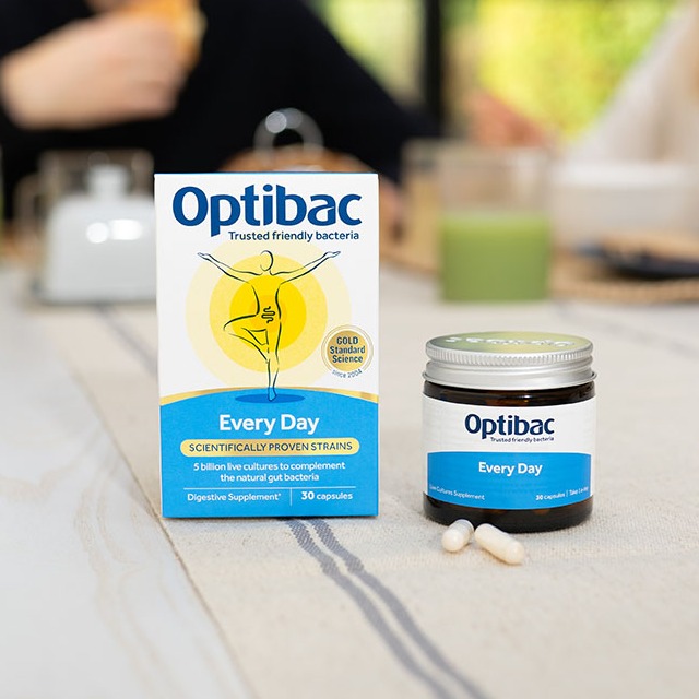 Optibac Every Day probiotic packaging and capsules on a table, with a blurred background of people and a green drink.