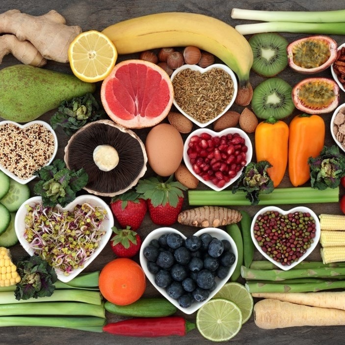 Assorted fruits, vegetables, and grains arranged with heart-shaped bowls of seeds, berries, and sprouts on a wooden surface.