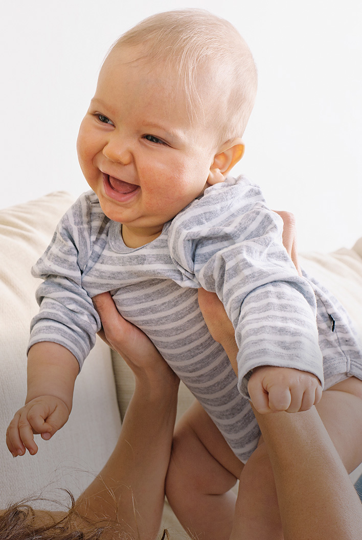 A happy baby in a striped onesie is lifted in the air, smiling joyfully with a white background.