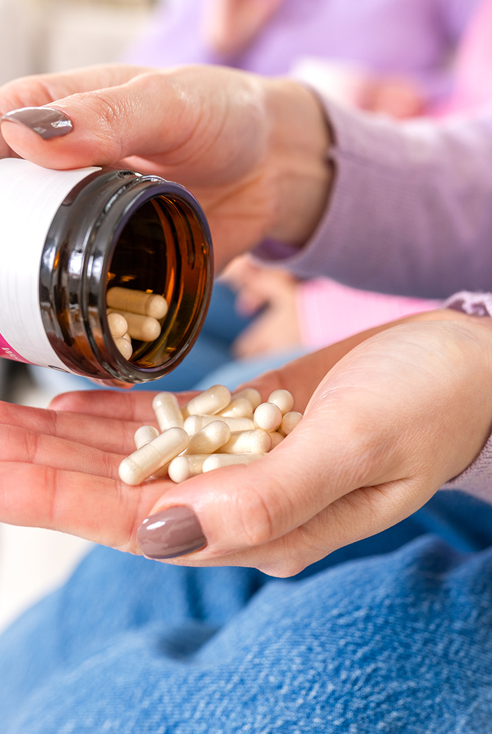 Person pouring white capsules from a brown bottle into their hand, wearing a purple sweater and jeans.
