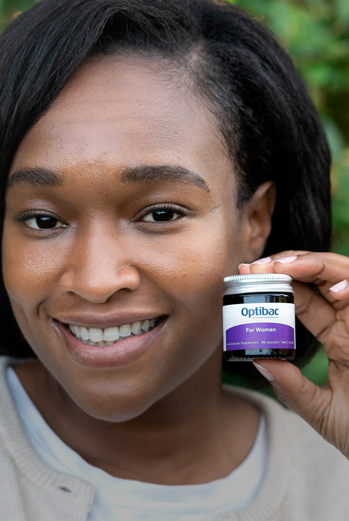 Smiling woman holding a small jar labeled "Optibac For Women" against a blurred green background.