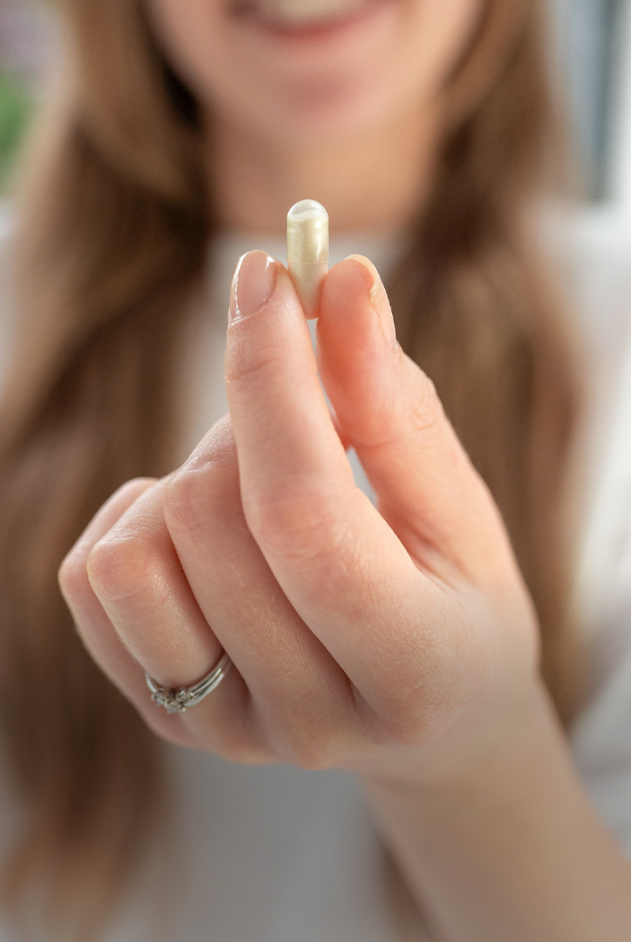 A person holding a small white capsule between their thumb and index finger, with a blurred background.