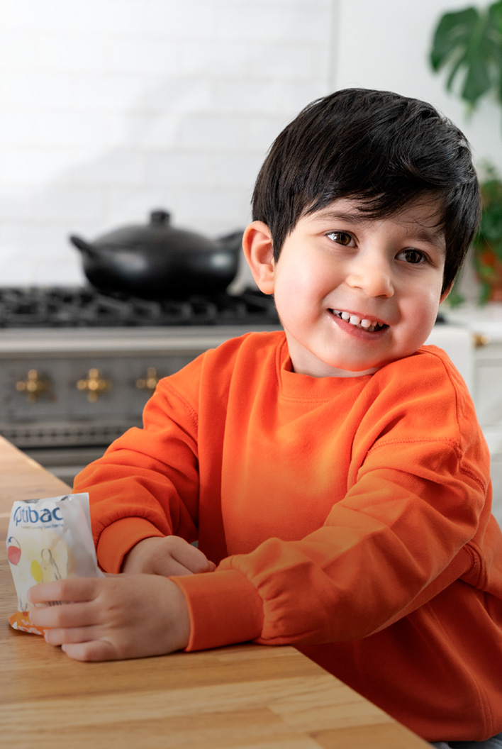Smiling child in an orange sweater holding a snack, standing by a kitchen counter with a stove and plant in the background.