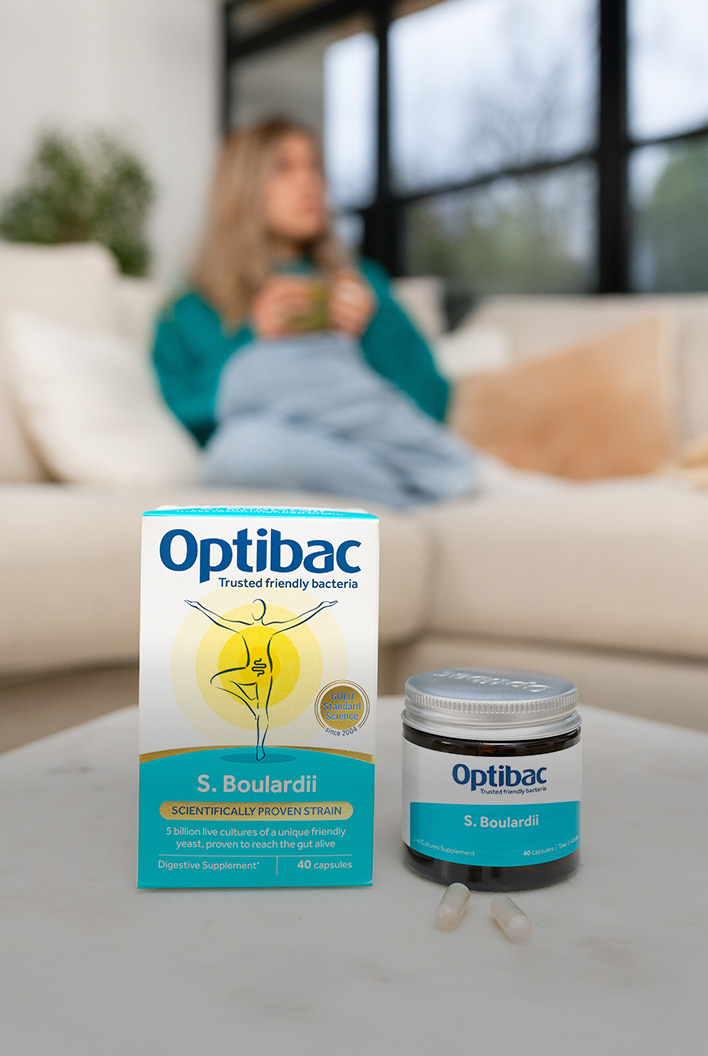 Optibac S. Boulardii probiotics on a table; a woman sits blurred in the background on a sofa.