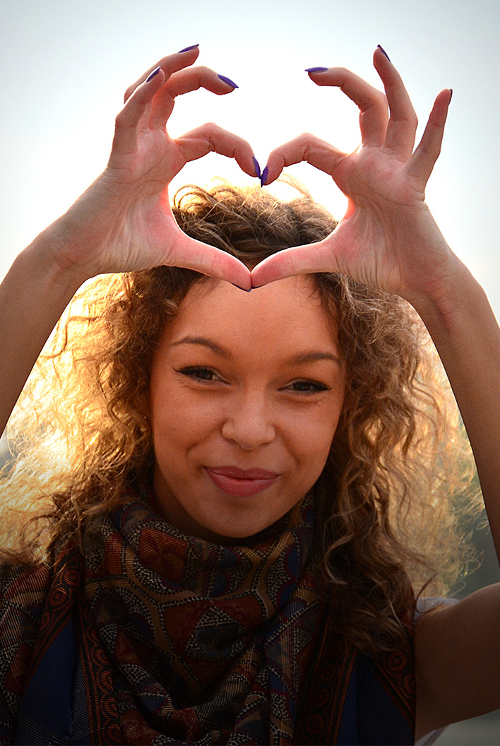Smiling woman with curly hair making a heart shape with her hands against a bright background.