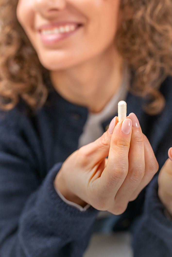 Smiling woman holding a small white capsule between her fingers, wearing a dark sweater.