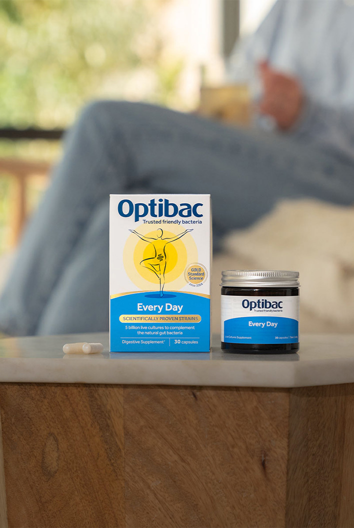 Optibac Every Day probiotics on a wooden table, with a person sitting in the background holding a cup.