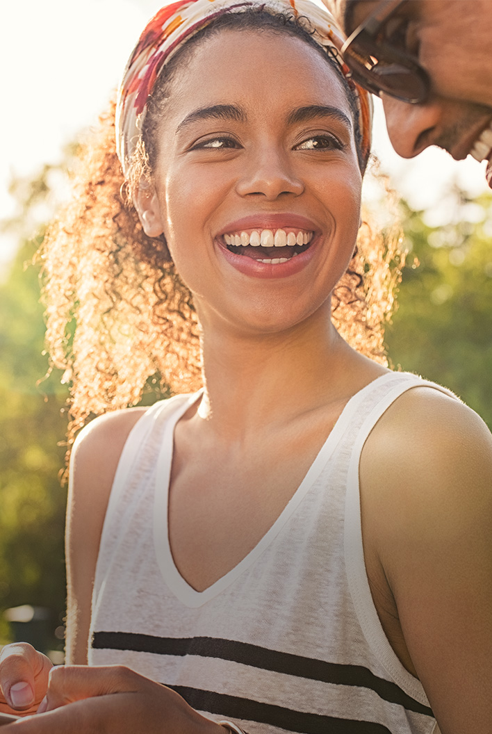 A smiling woman with curly hair and a headband stands outdoors in the sunlight, wearing a sleeveless top and looking at someone beside her.