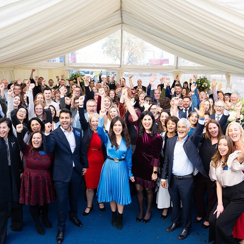 A large group of people in a celebratory pose under a white tent, smiling and raising their fists, with a blue carpet and floral decorations.