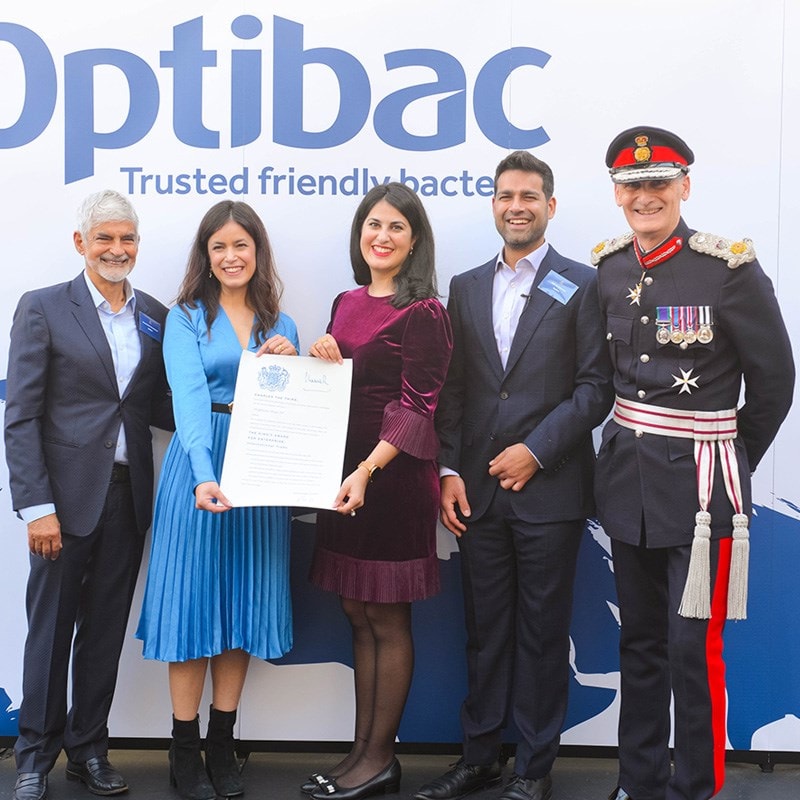 Five people smiling, standing in front of an "Optibac" backdrop. One holds a certificate. A man in a ceremonial uniform is on the right.