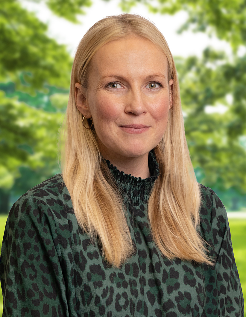 A woman with long blonde hair, wearing a green leopard print top, smiles slightly against a blurred background of greenery.