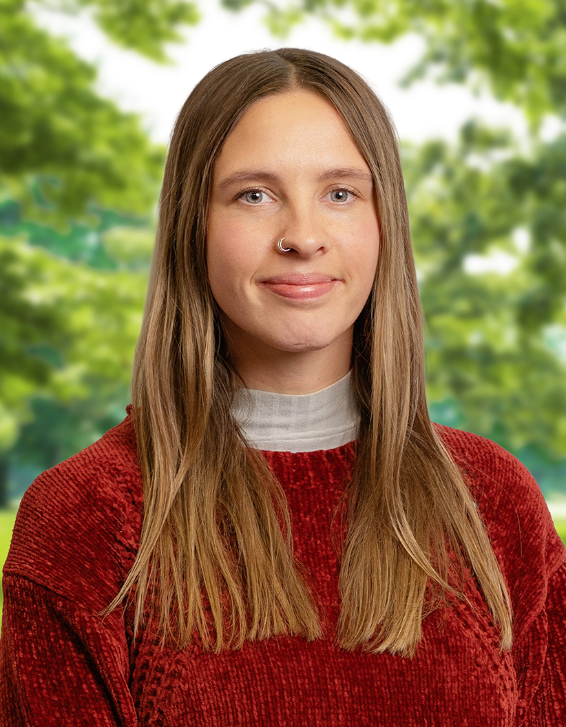 A person with long brown hair and a nose ring, wearing a red sweater, stands outdoors with green trees blurred in the background.