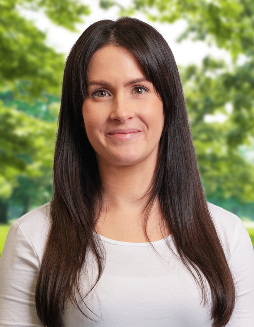 Smiling woman with long dark hair in a white shirt, standing outdoors with blurred green trees in the background.