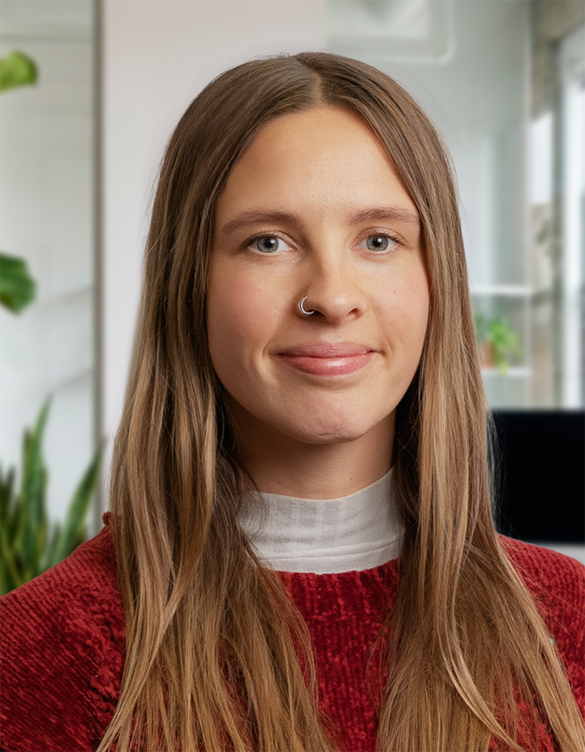 A woman with long brown hair and a nose ring smiles slightly, wearing a red sweater and white top, in a bright indoor setting with plants.