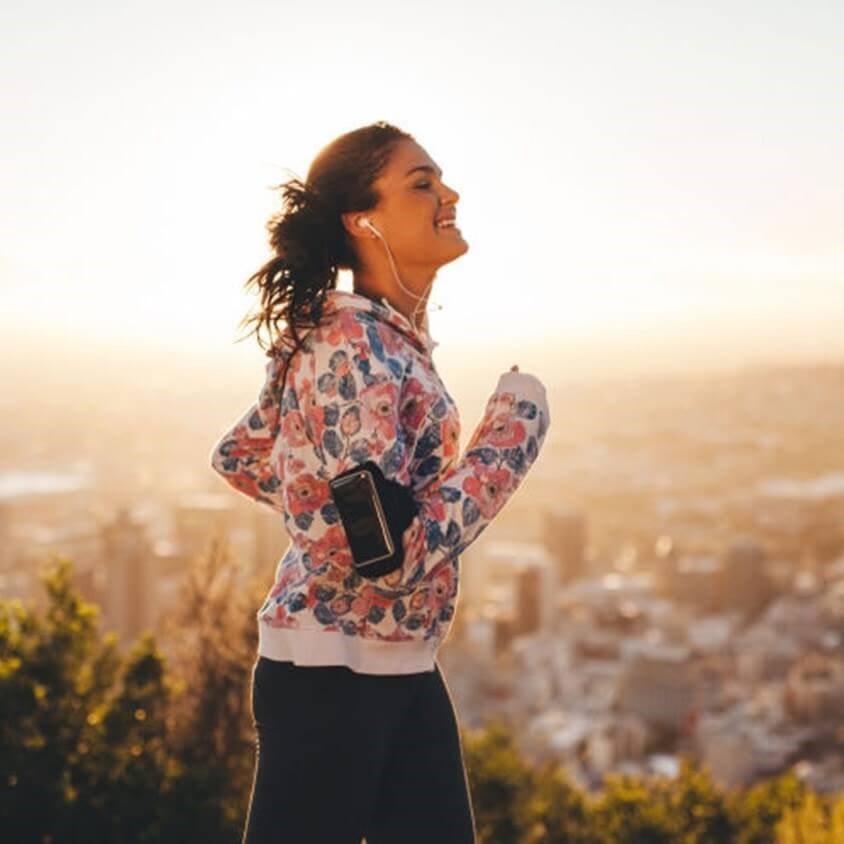 Woman jogging with earphones, wearing a floral hoodie, smiles against a cityscape and sunrise background.