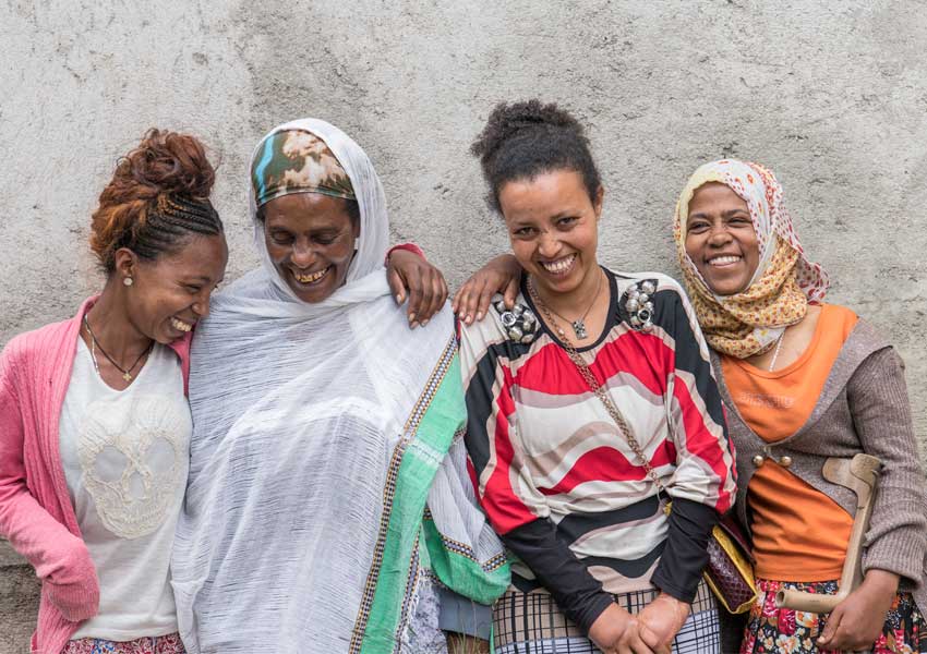Four women standing close together, smiling and laughing against a textured wall. They wear colorful, varied clothing and headscarves.