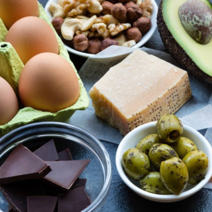A variety of foods including eggs, walnuts, hazelnuts, avocado, cheese, green olives, and dark chocolate arranged on a dark surface.