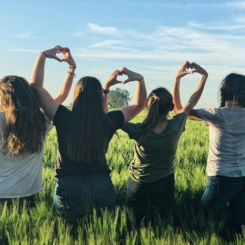 Four people standing in a green field, facing away, forming heart shapes with their hands against a blue sky.