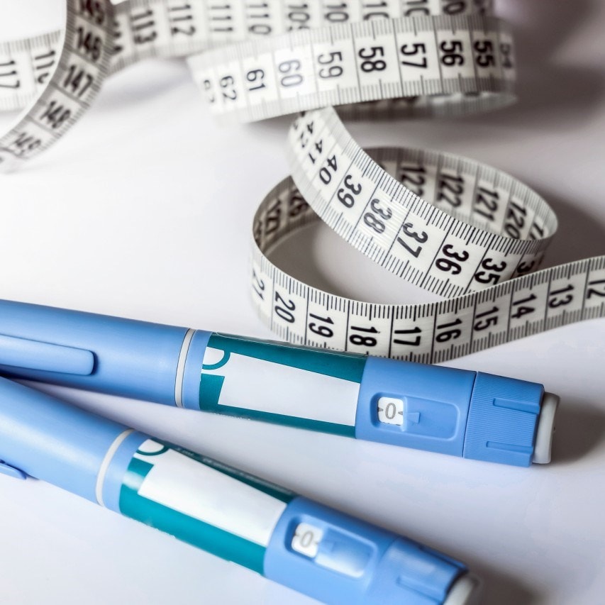 Two blue insulin pens lie on a surface beside a coiled white measuring tape with black numbers.