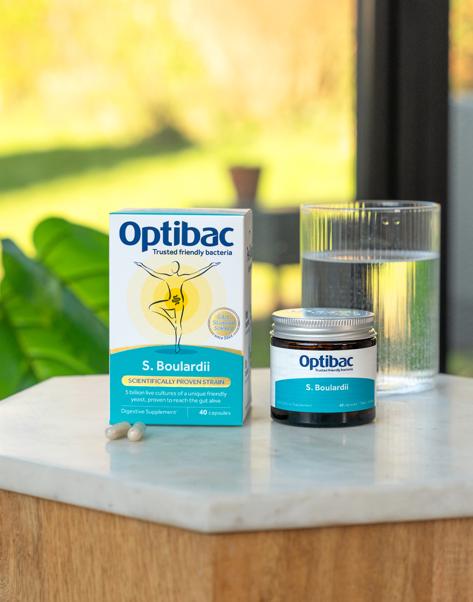 Optibac S. Boulardii probiotic box and jar on a table with two capsules and a glass of water, set against a blurred natural background.