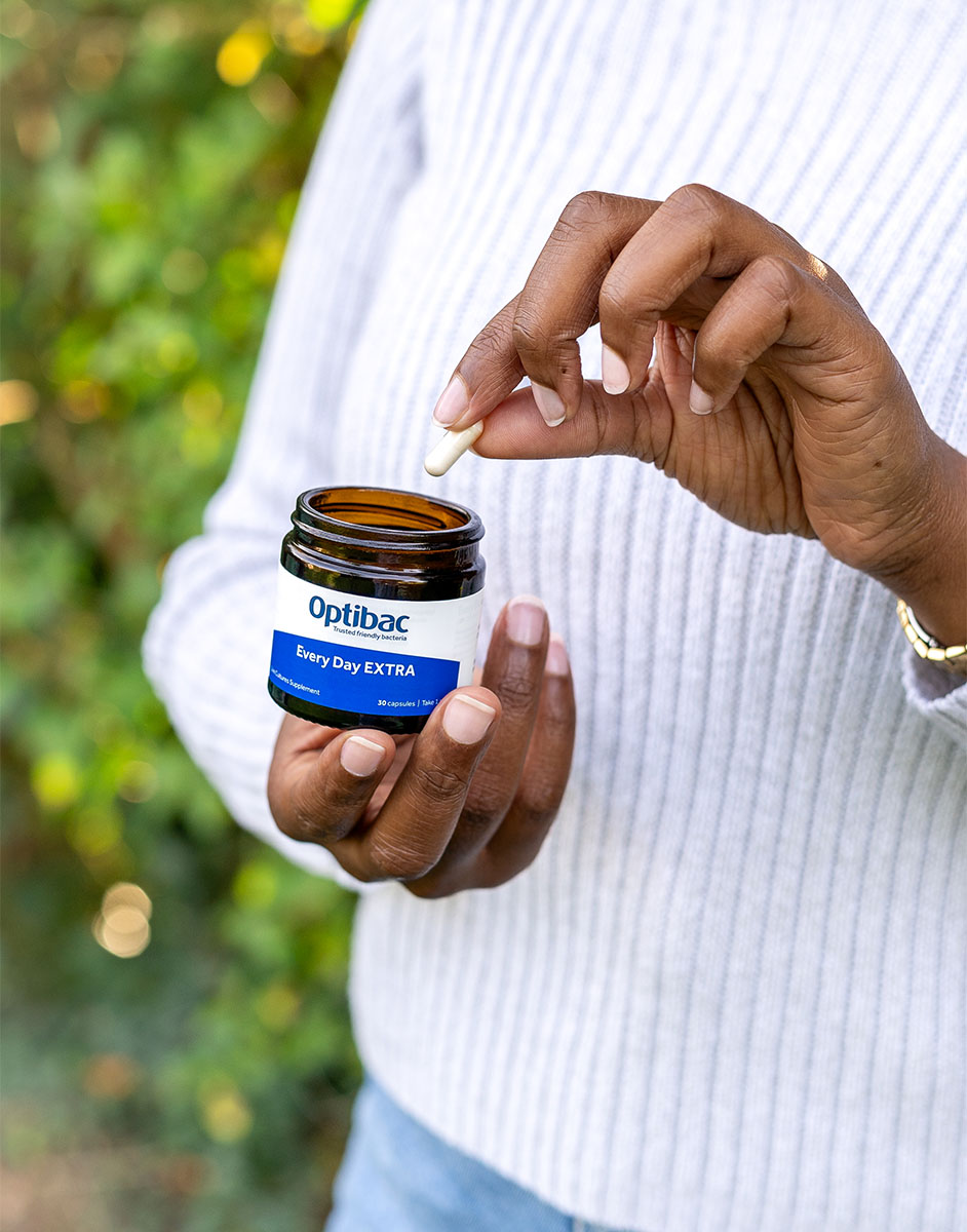 Person holding an Optibac Every Day EXTRA supplement jar, taking a capsule. Blurred green foliage in the background.