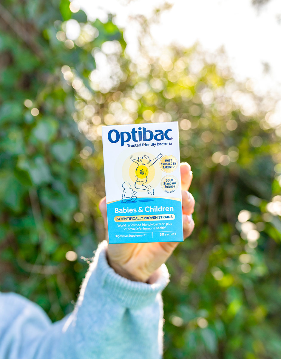 A hand holds a box of Optibac probiotics for babies and children against a blurred outdoor background of greenery.
