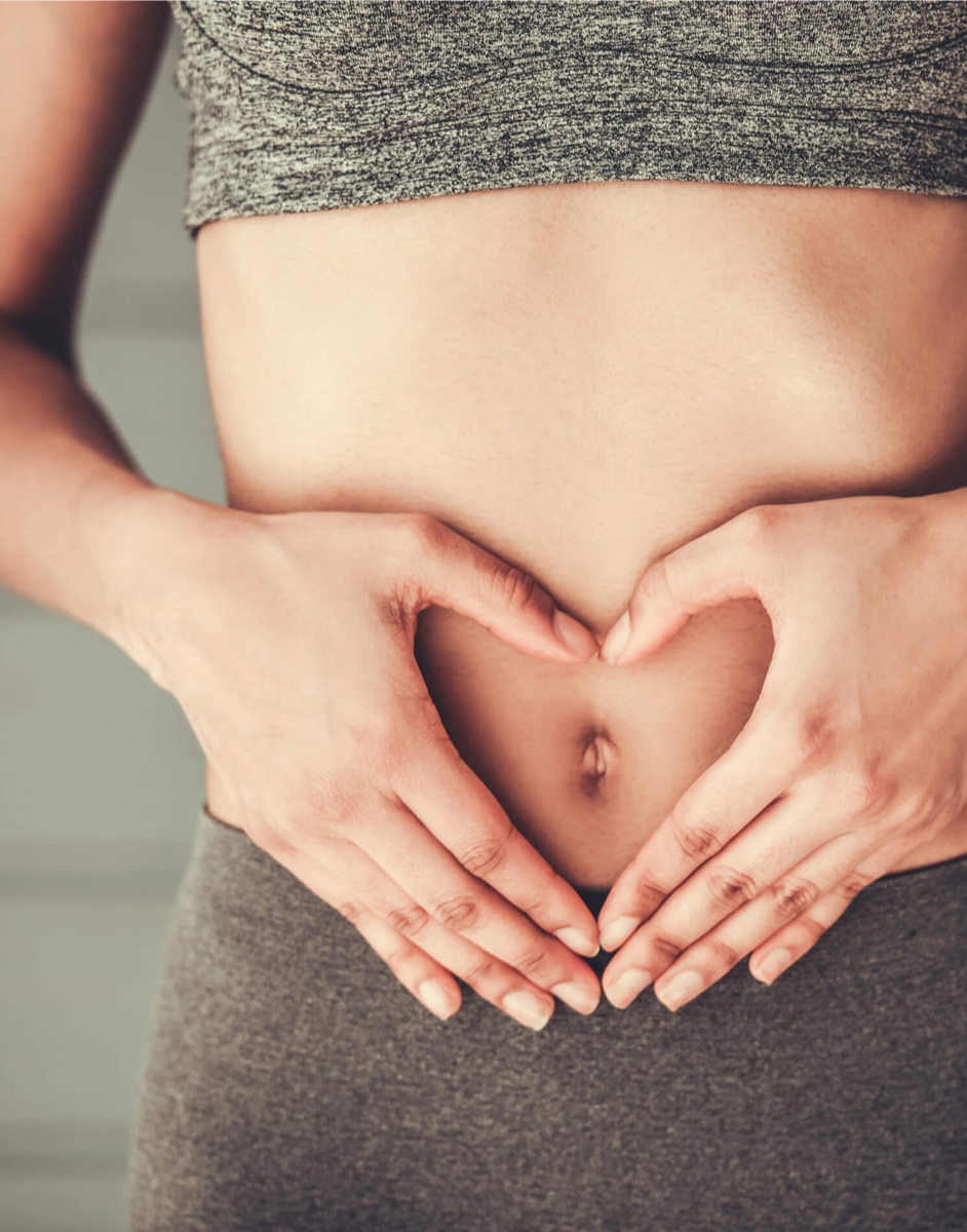 Person in a grey outfit forming a heart shape with hands over their abdomen, symbolising care or love for health and wellness.