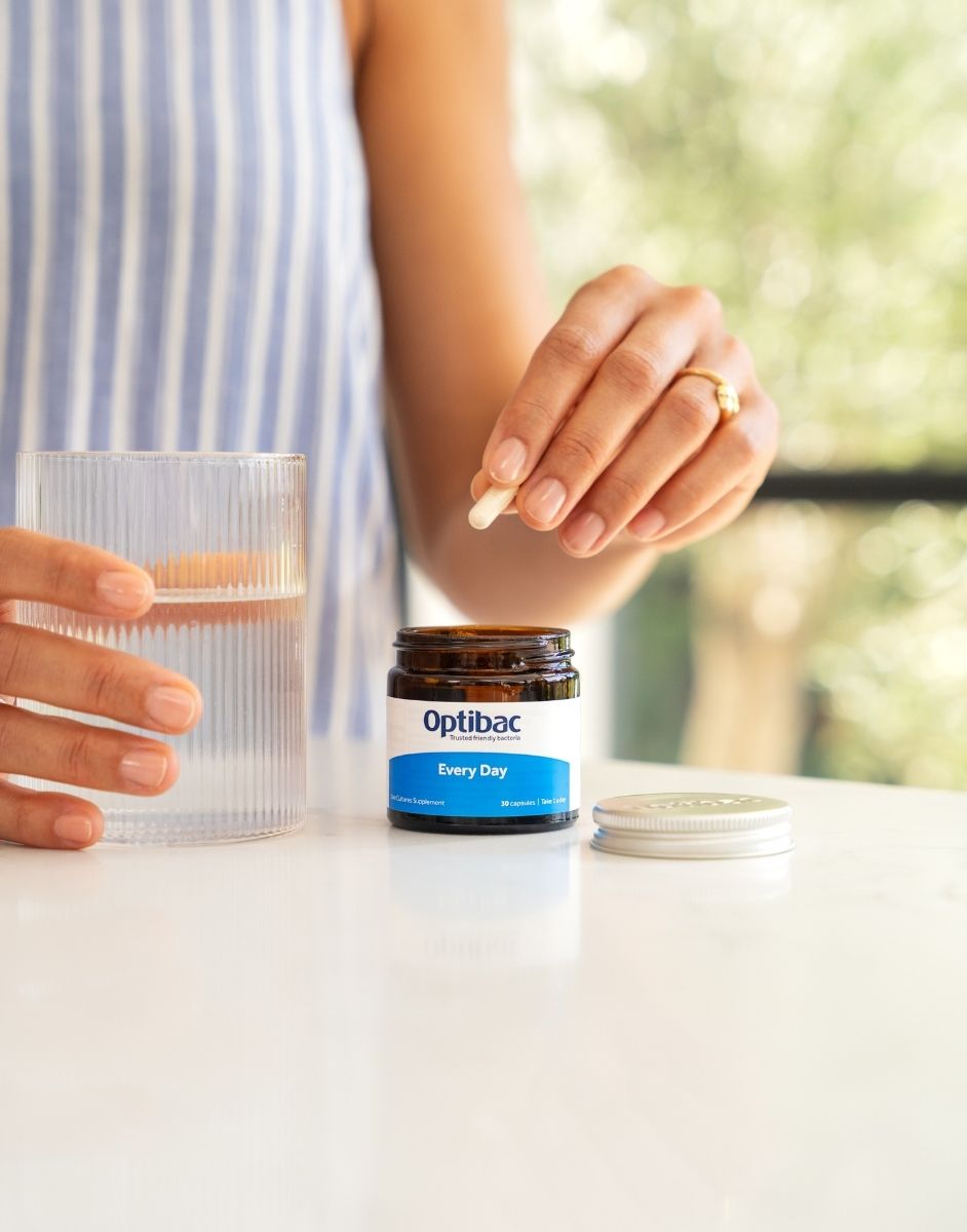 A person is holding a capsule over an open Optibac "Every Day" supplement jar, with a glass of water on a white countertop.