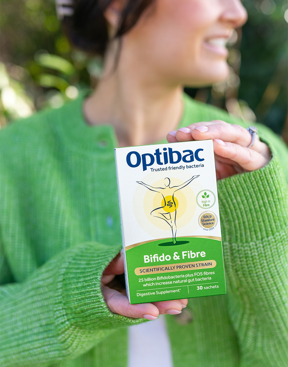 Person in a green sweater holding a box of Optibac Bifido & Fibre supplement, smiling outdoors.
