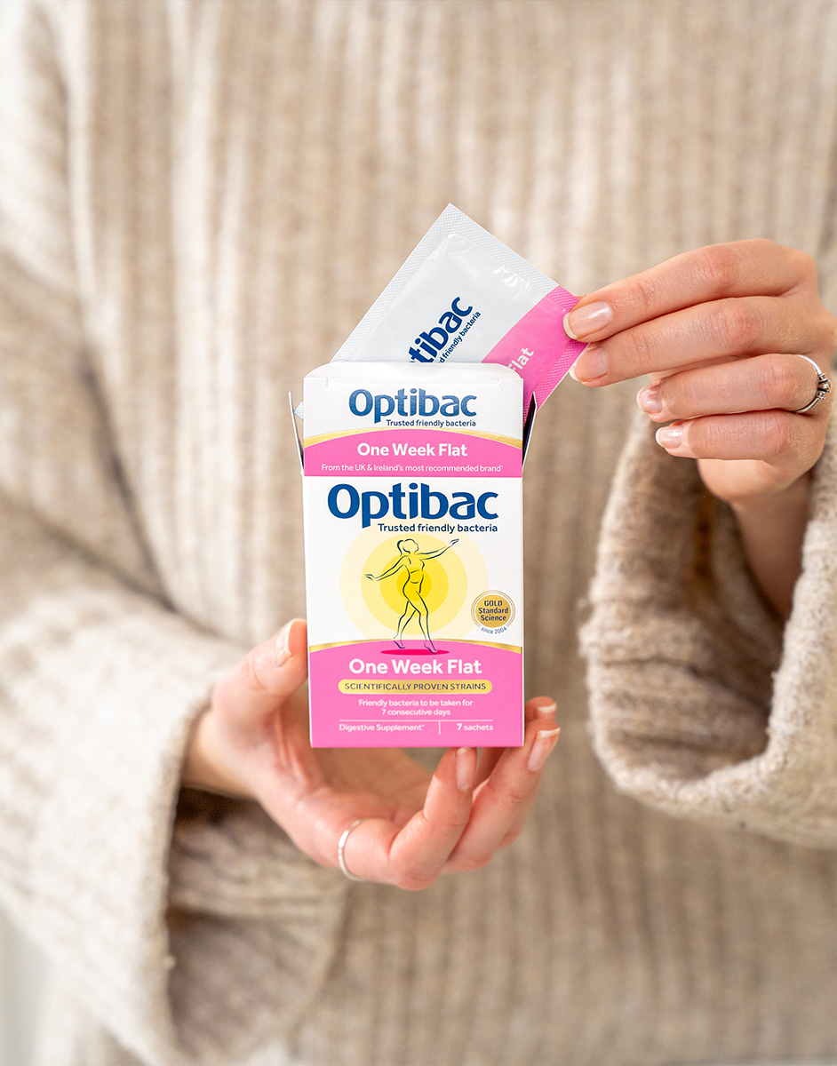 Person holding an Optibac One Week Flat probiotic box and sachet, wearing a beige knitted sweater.