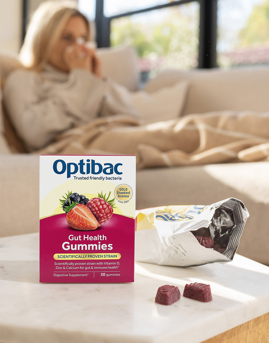 Box of Optibac Gut Health Gummies on a table with a woman in the background sitting on a couch, wrapped in a blanket.