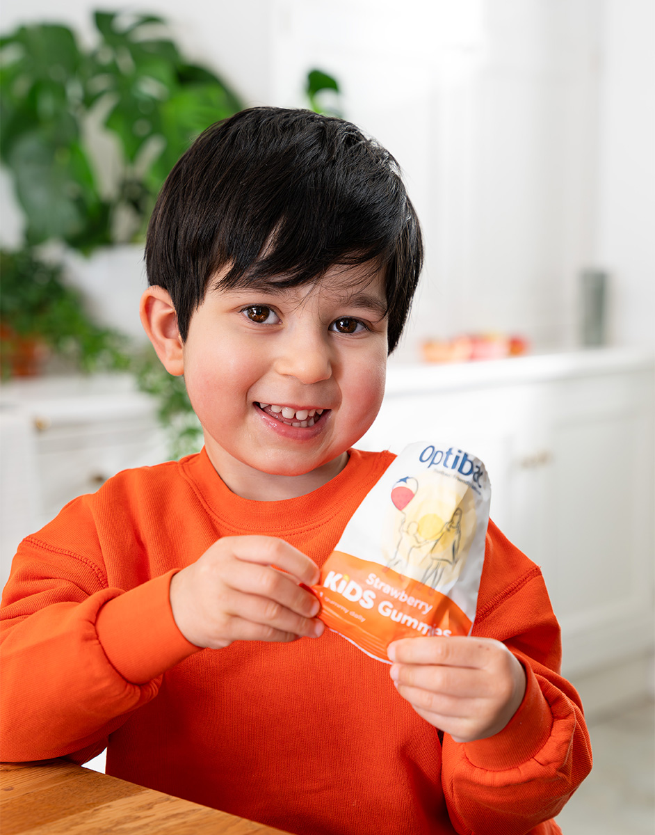 Smiling child in an orange sweater holds a pack of strawberry-flavored kids gummies in a bright kitchen.
