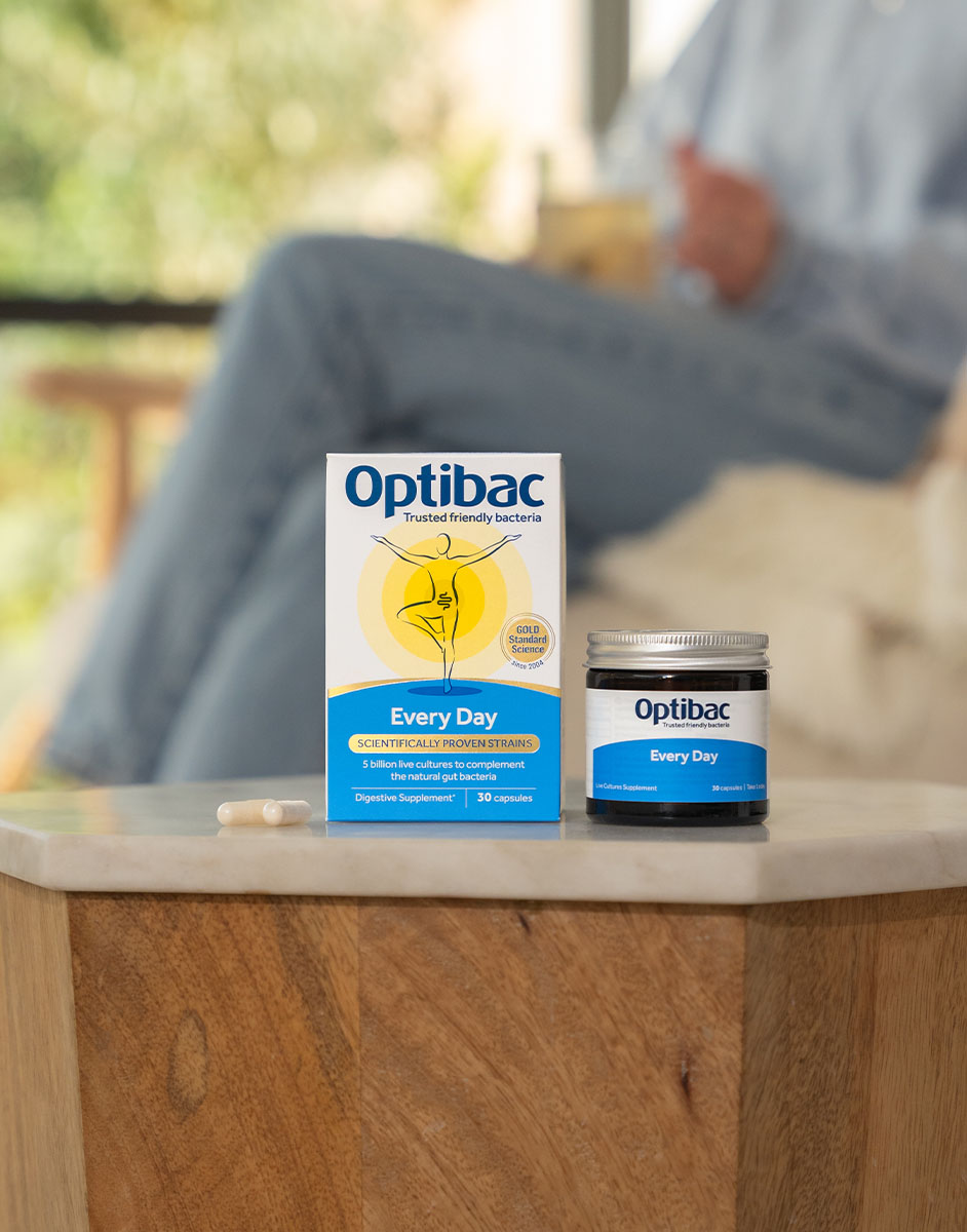 Optibac probiotics box and jar on a wooden table with a capsule beside them; person sitting blurred in the background.