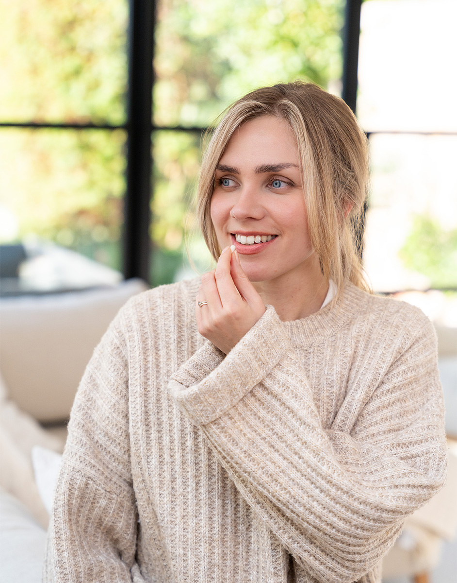 A smiling woman with blonde hair in a cozy beige sweater stands indoors, with a blurred background of windows and greenery.