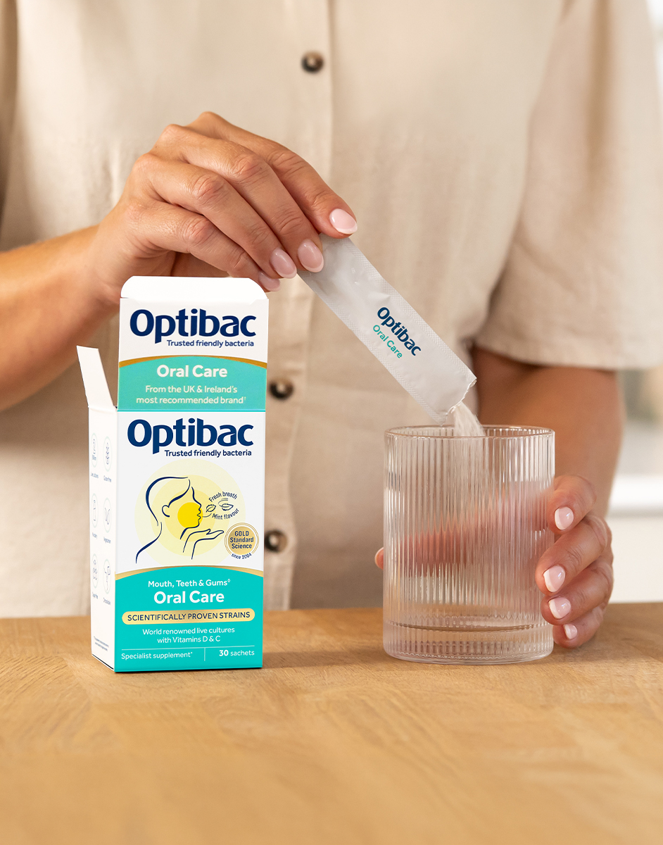 A person pours Optibac Oral Care powder into a clear glass next to an open box of the product on a wooden table.