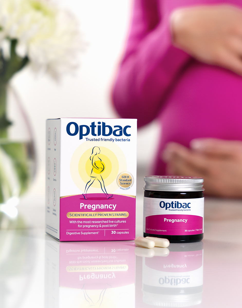 Optibac Pregnancy supplement packaging and capsules on a reflective surface, with a blurred background of a person in pink holding their belly.