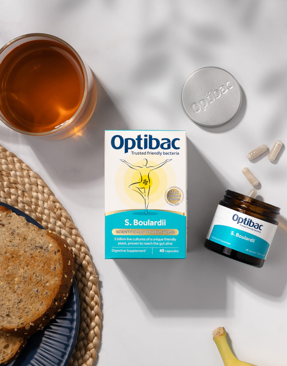 Optibac S. Boulardii supplement box with capsules, a glass of tea, bread on a woven mat, and a banana on a white surface.