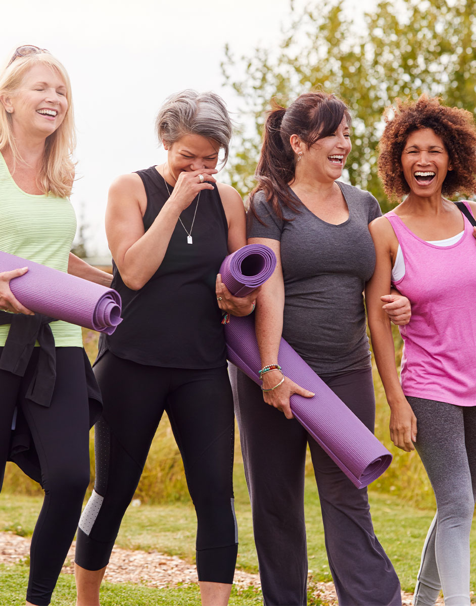 Four women laughing and holding yoga mats while walking outdoors in casual athletic wear, surrounded by greenery.