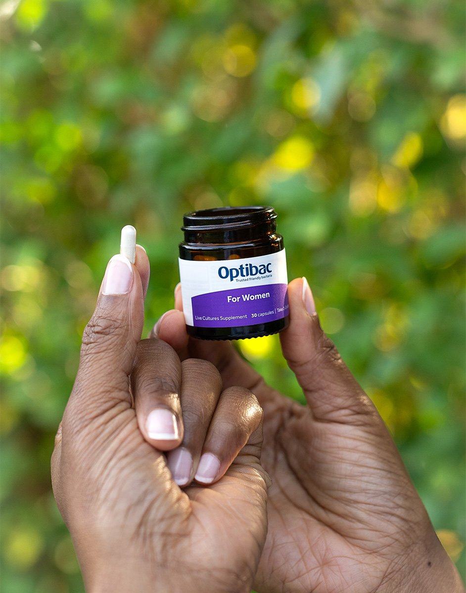 A person holds a small white capsule and a jar labeled "Optibac For Women" against a blurred green background.