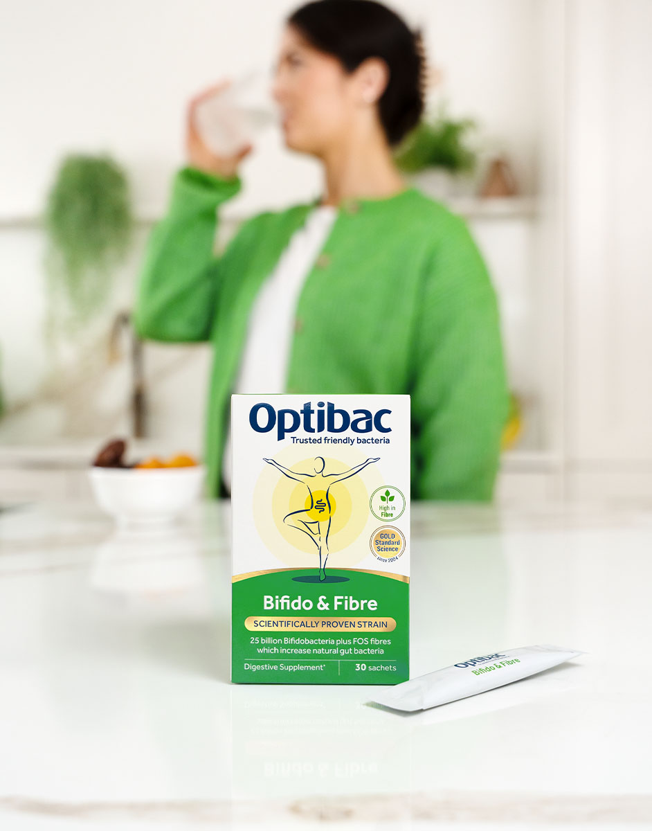 Box of Optibac Bifido & Fibre supplement on a counter, with a person in a green jacket drinking from a glass in the background.