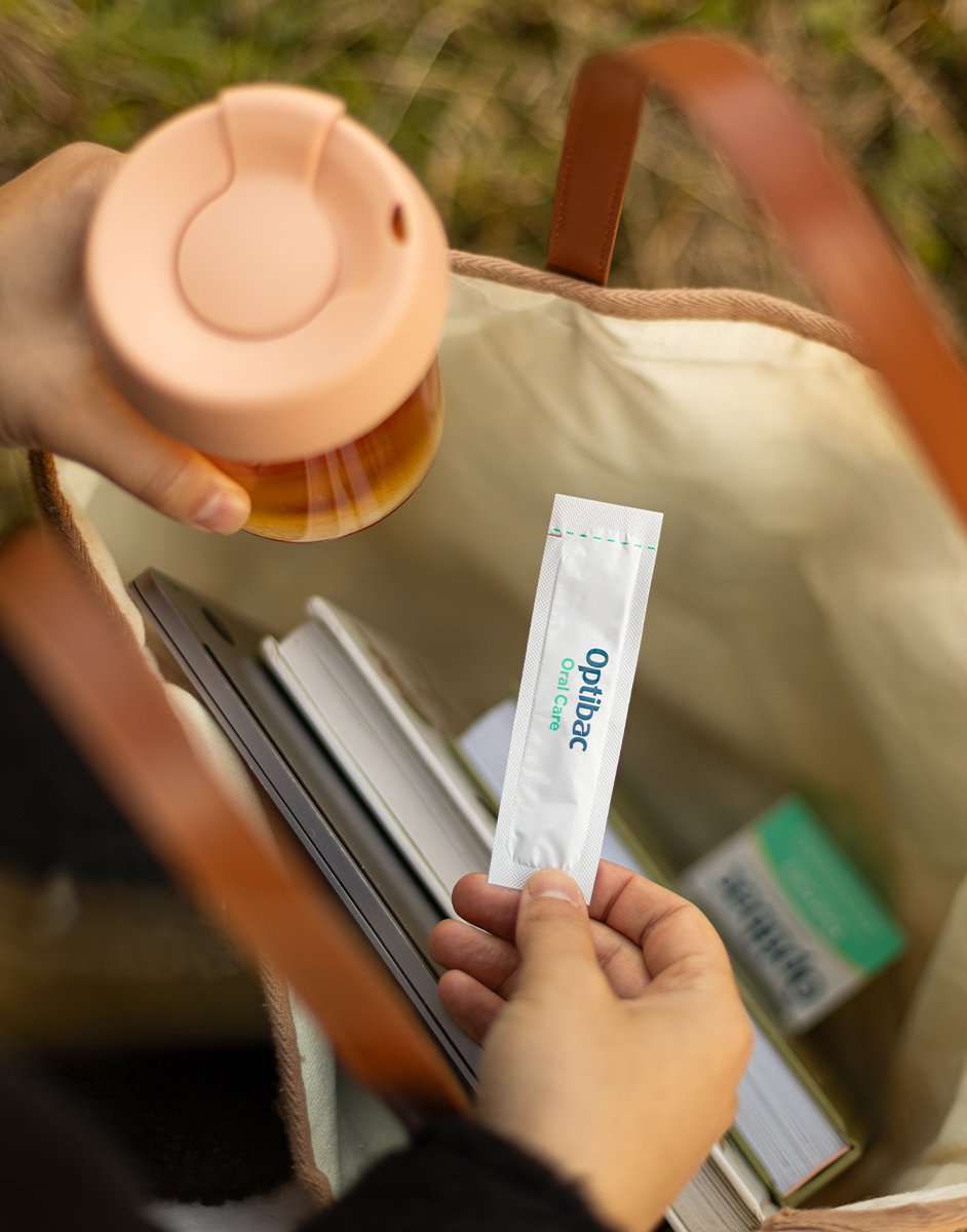 A person holds a travel mug and is pulling a sachet labeled "Optibac Oral Care" from an open bag containing books and a box.