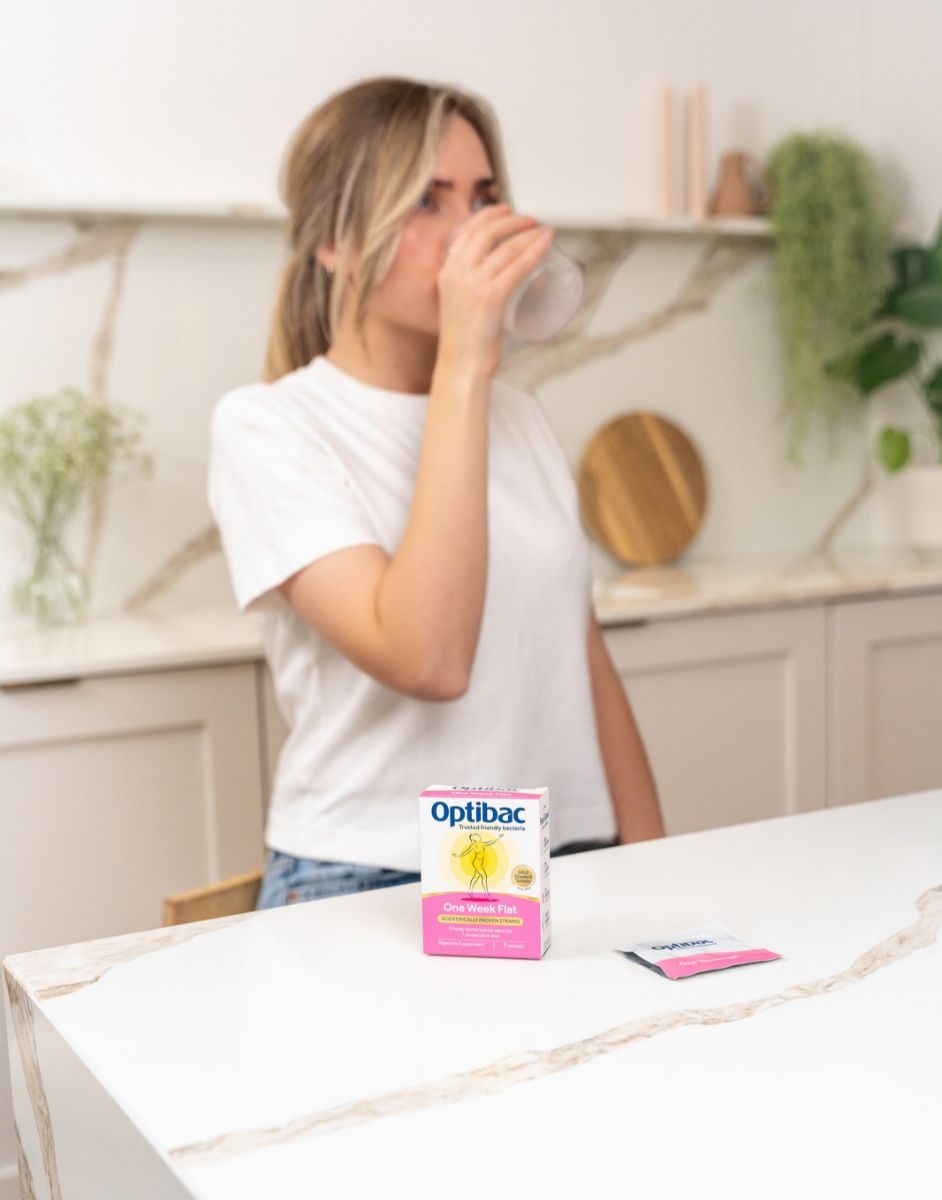A person in a kitchen drinks from a glass, with Optibac One Week Flat probiotic product on the countertop.