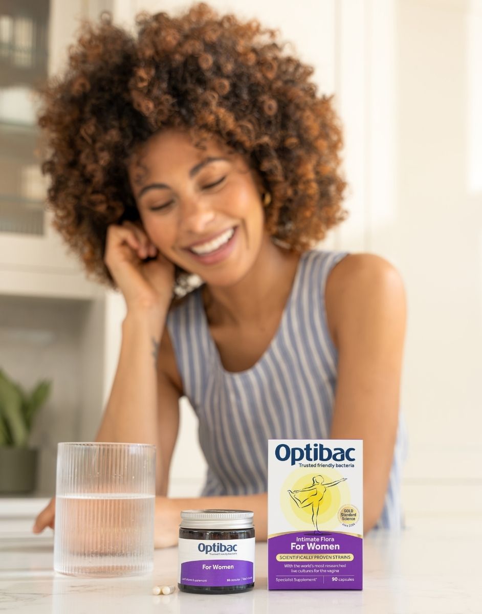 Woman smiling with Optibac supplements on a table, featuring a glass of water and product packaging focused on women's health.
