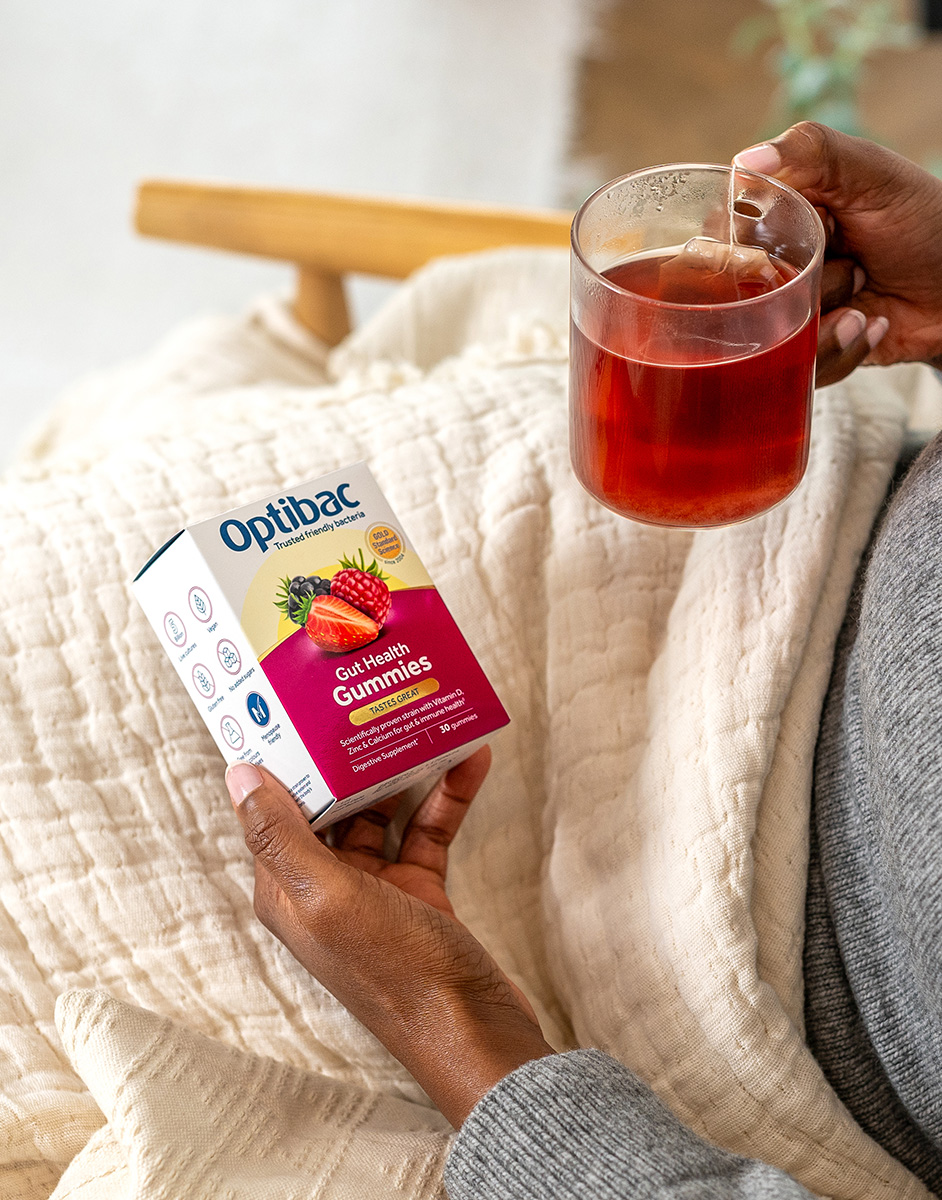 Person holding Optibac Gut Health Gummies box and a glass of red drink, sitting on a beige blanket.