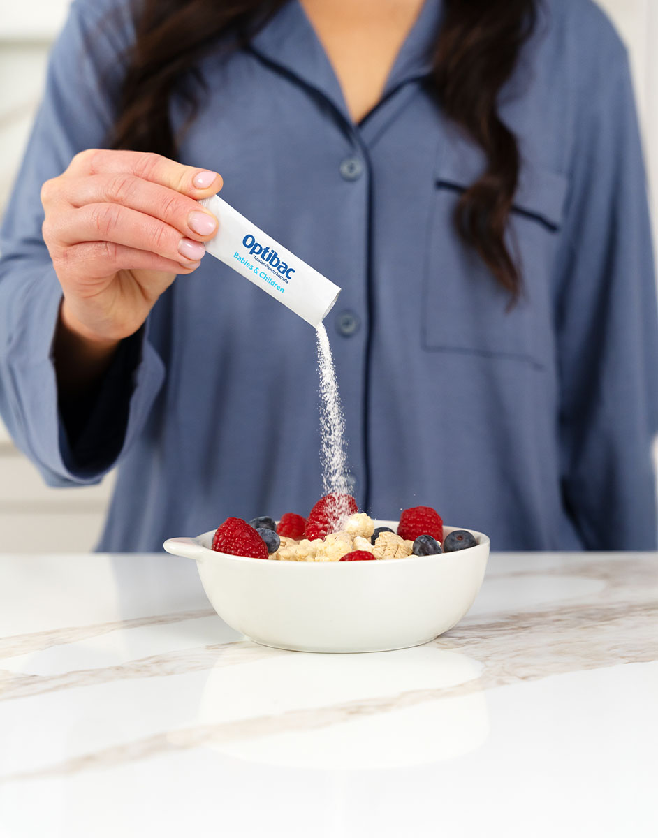 Person sprinkling Optibac powder over a bowl of oatmeal topped with raspberries, blueberries, and nuts on a marble countertop.